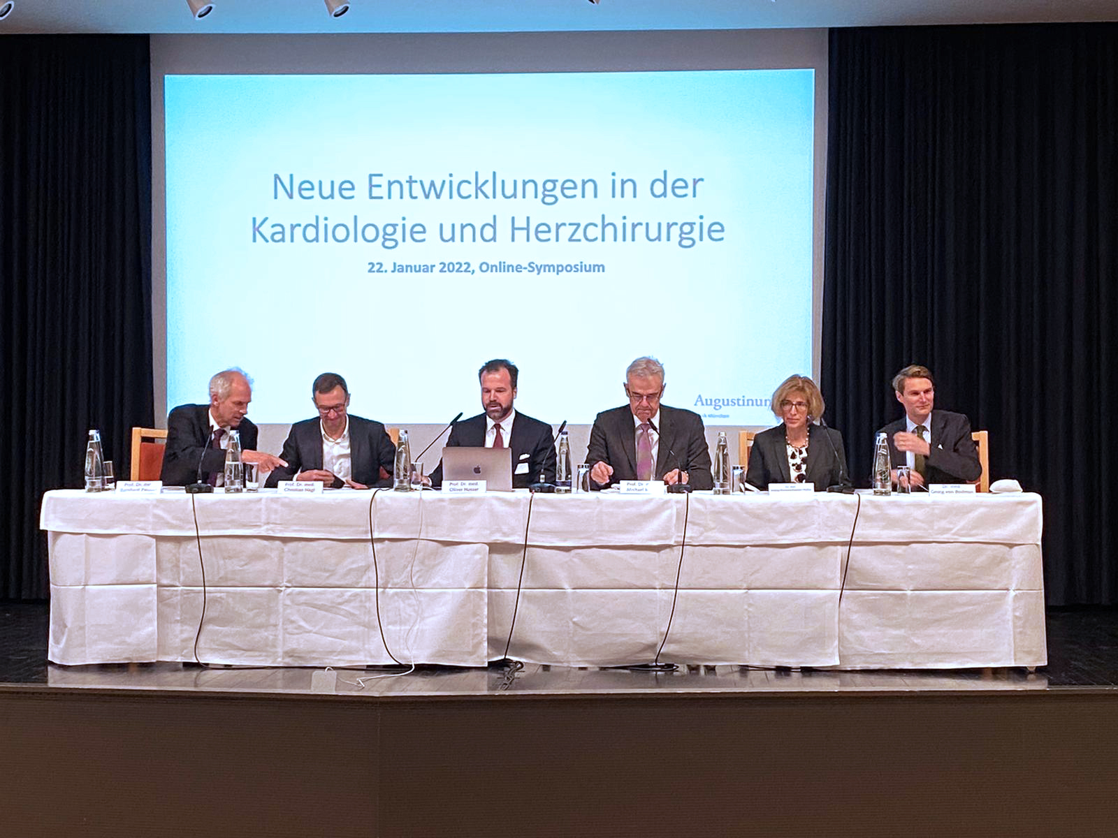 Fünf Ärzte und eine Ärztin sitzen auf einem Podium hinter einem Tisch. Im Hintergrund wird der Titel des Symposiums projiziert.
