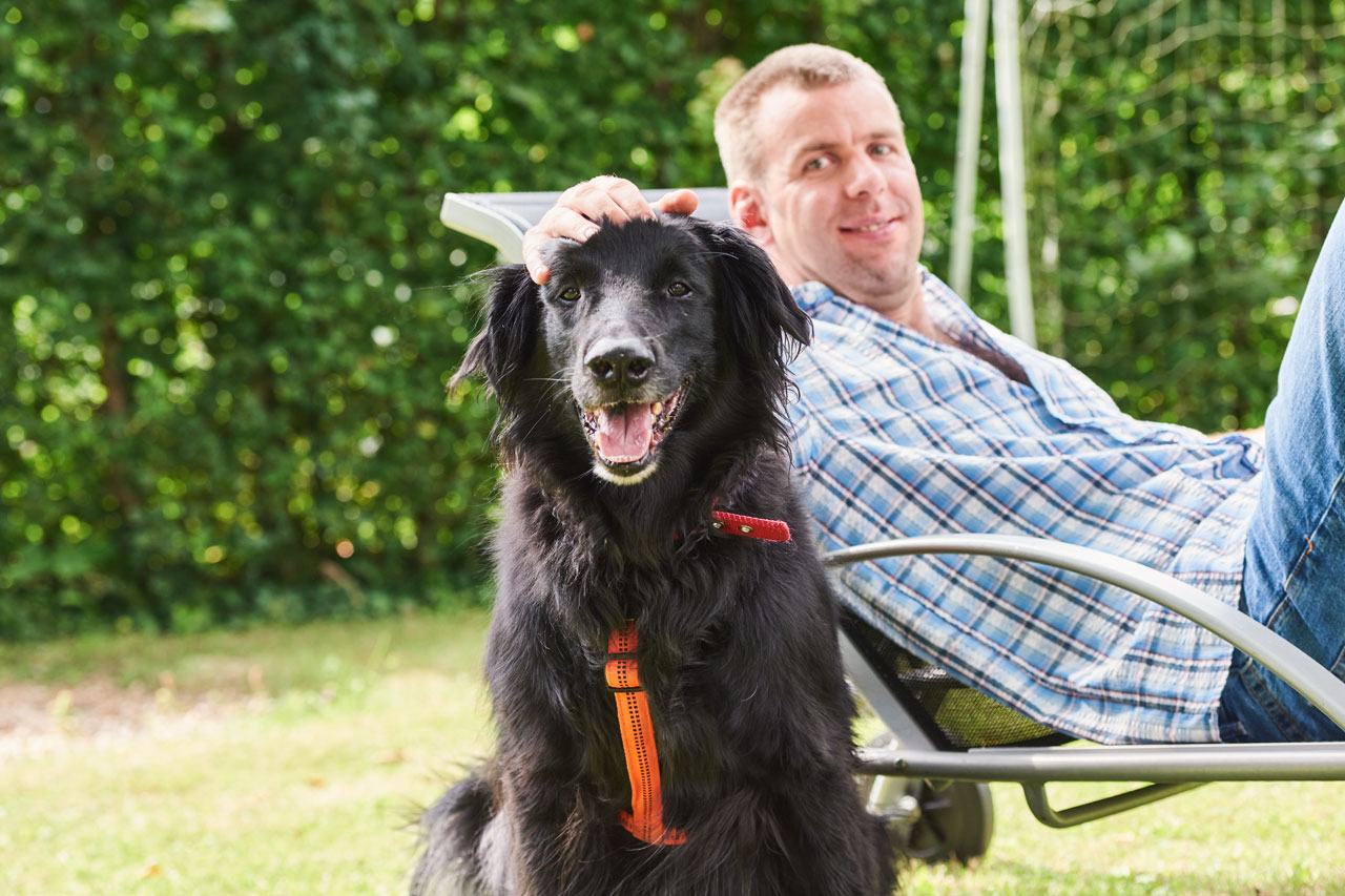 Ein Mann liegt auf einer Liege im Garten, davor sitzt ein Hund