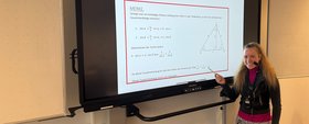 Eine Lehrerin, die ein Headset trägt, zeigt lächelnd auf den Bildschirm neben ihr, auf dem Lerninhalte angezeigt werden