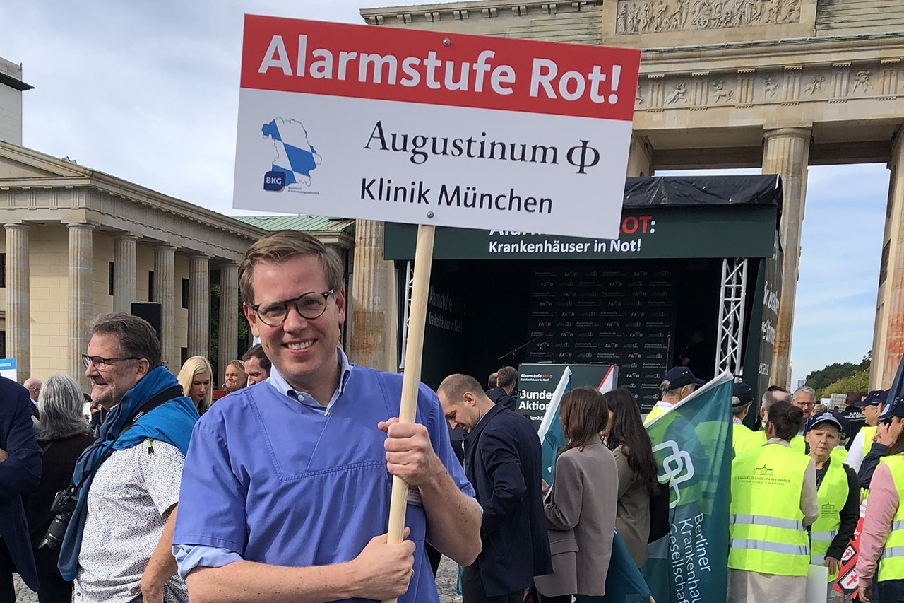 Ein Mitarbeiter der Augustinum Klinik hält ein Schild mit der Aufschrift "Alarmstufe Rot! Augustinum Klinik München"