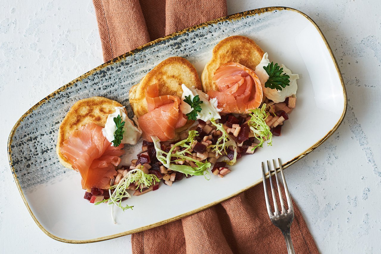 Auf einem ovalen Teller mit grauem Rand am oberen Ende sind drei Blinis mit drei Scheiben Lachs in einer Reihe angerichtet. Darunter, ebenfalls in einer Linie, Salat mit Apfel und Rote Beete.