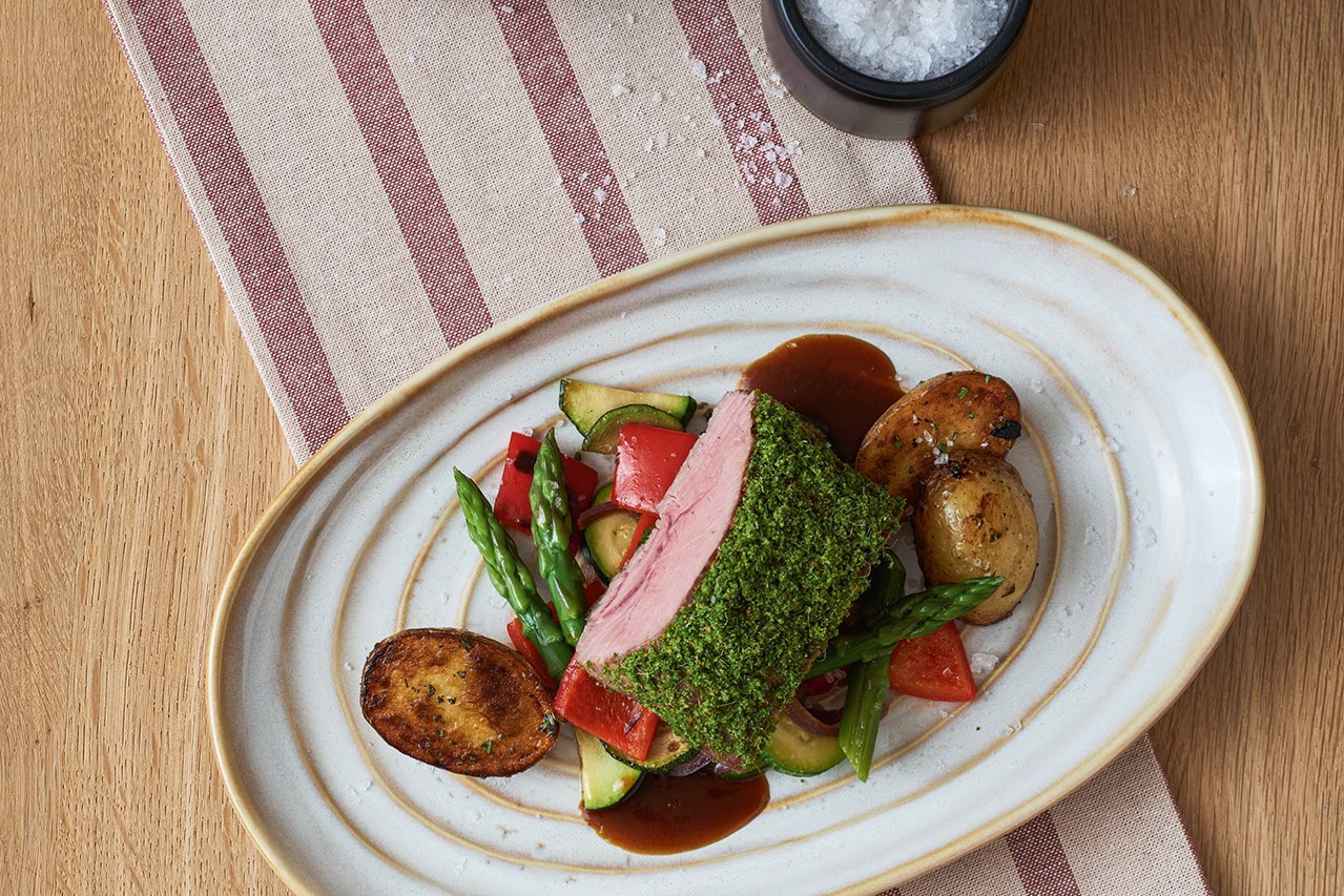 Auf einem runden ovalen Teller liegt ein rosa gebratenes Stück Lamm mit grasgrüner Kruste. Darunter buntes Frühlingsgemüse (Paprika, Zucchini, grüner Spargel) und gebratene Kartoffelhälften.
