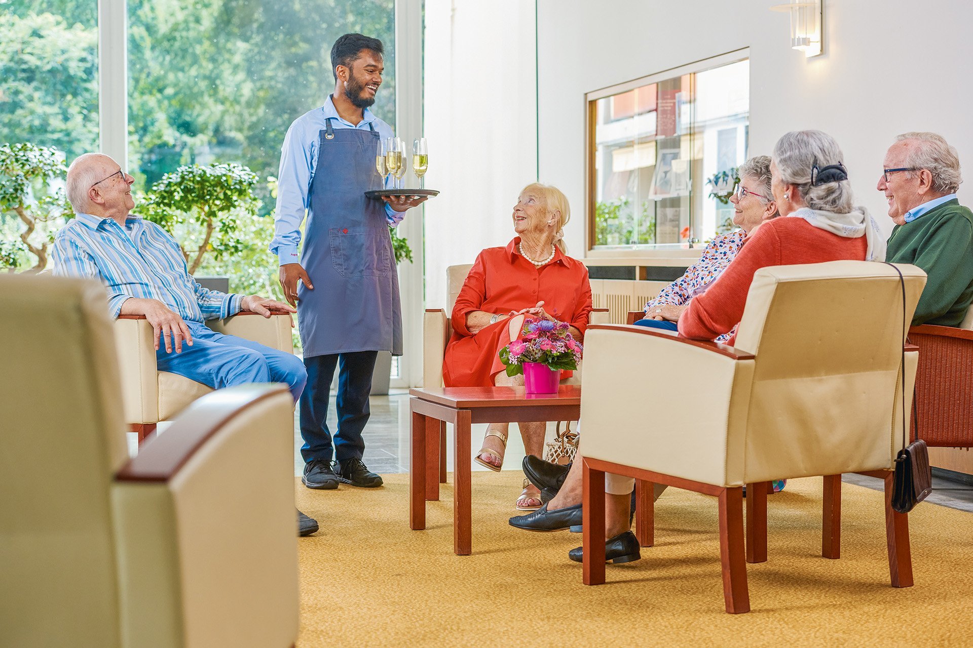Eine Gruppen von Senior*innen sitzt gemeinsam im Foyer und ein Mitarbeiter des Augustinum bringt ihnen Sektgläser