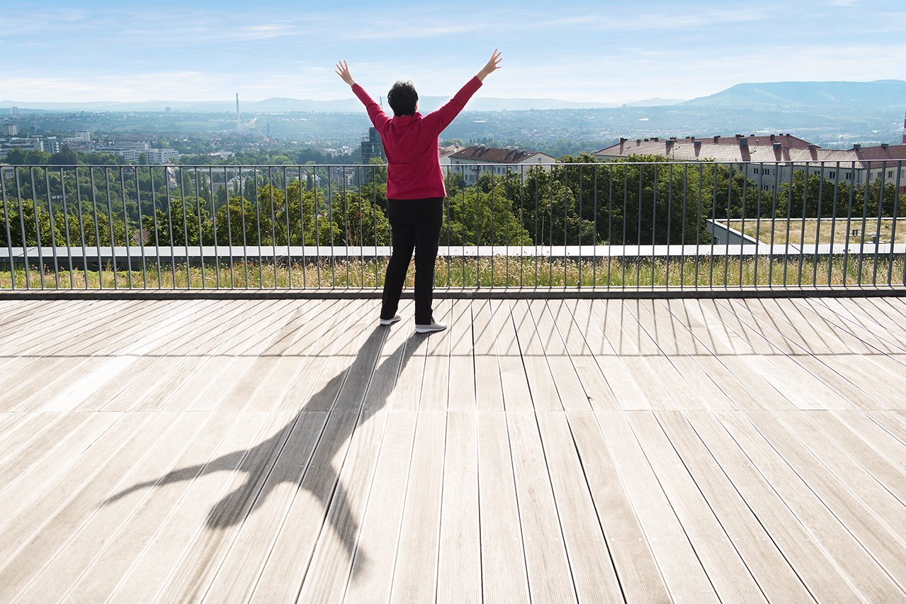 Ein Seniorin steht mit nach oben gestreckten Armen auf einer Terrasse 