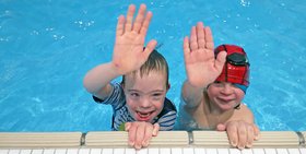 zwei Kinder mit Badekappe sind am Rand eines Schwimmbeckens und halten Ihre Hand zum Einschlagen hin