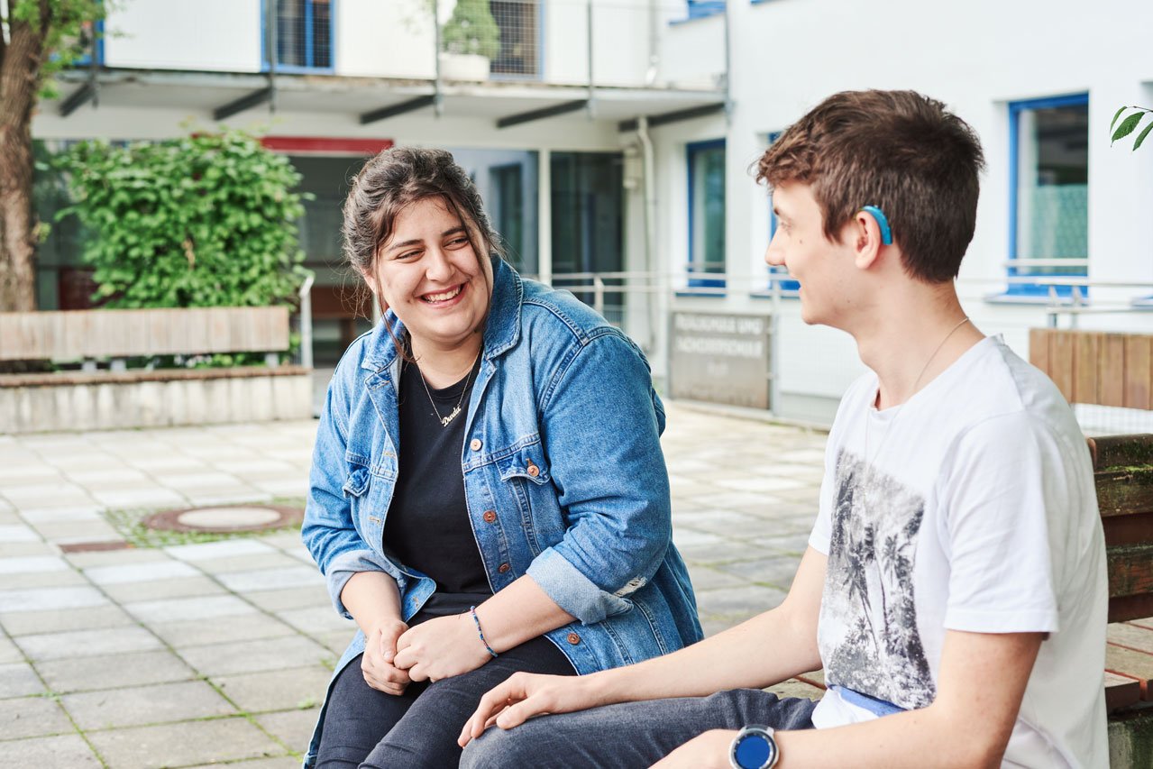 Zwei Schüler*innen sitzen auf einer Bank im Pausenhof und unterhalten sich