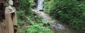 Augustinum Roth Regentrude Skulptur der Regentrude vor einem Wasserfall mit Bachlauf