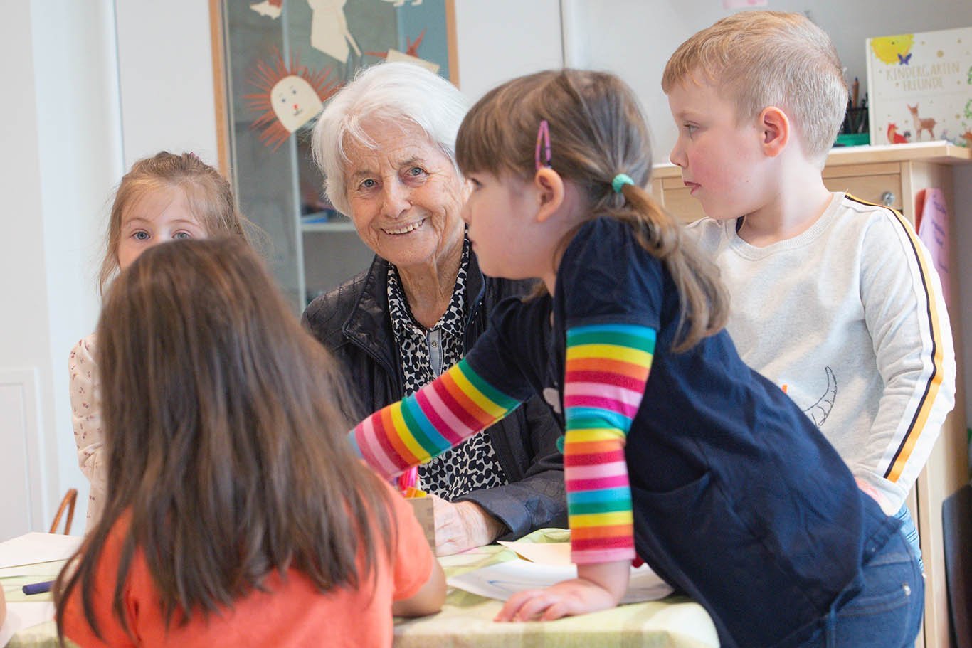 Seniorin an einem Tisch umringt von vier Kindergartenkindern