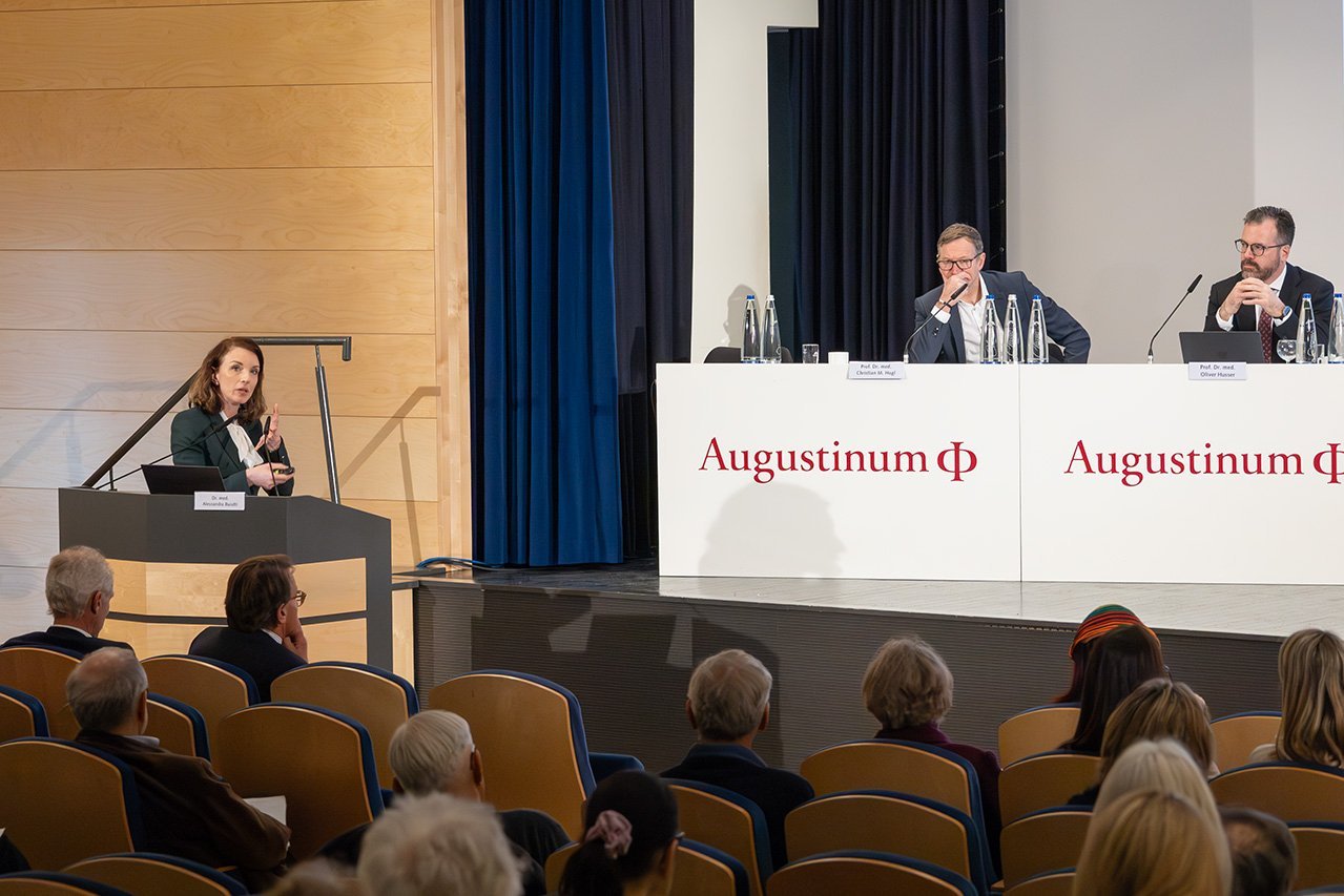 Eine Oberärztin der Augustinum Klinik, Dr. Buiatti, spricht zum Publikum, im Hintergrund sitzen zwei Veranstalter auf der Bühne