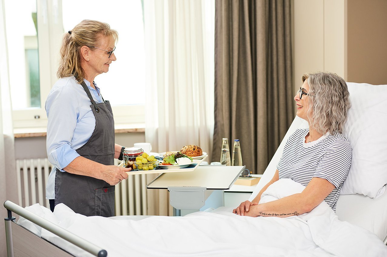 Eine Mitarbeiterin der Augustinum Klinik bringt einer im Bett liegenden Patientin ihr Essen 