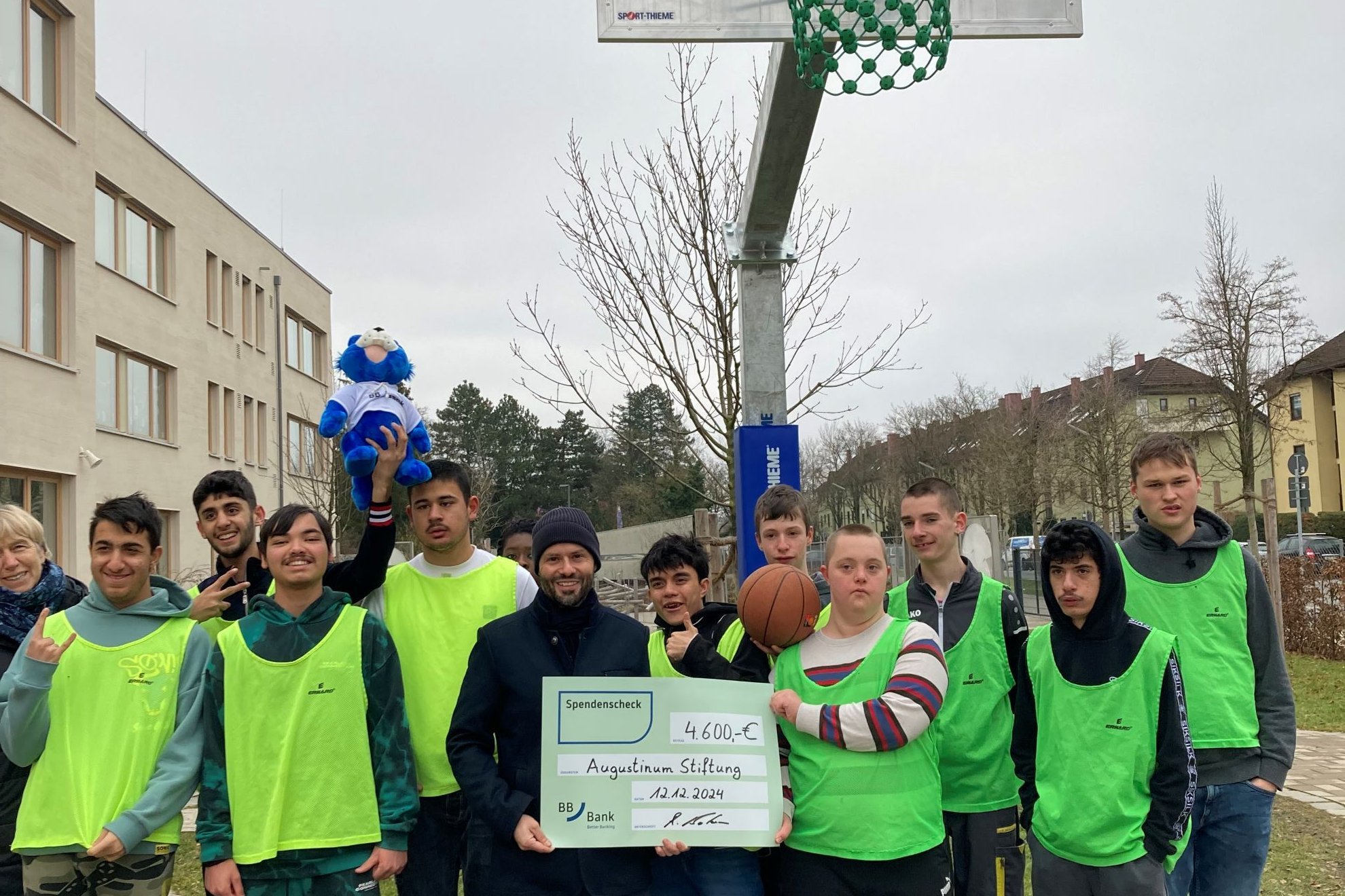 Schüler*innen vor Basketballkorb und Mann von BBBank, der Spendenscheck in den Händen hält, stehen vor einem Basketballkorb im Pausenhof