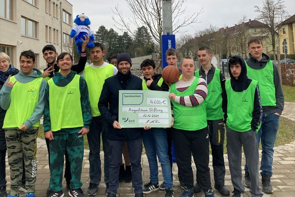 Schüler*innen vor Basketballkorb und Mann von BBBank, der Spendenscheck in den Händen hält, stehen vor einem Basketballkorb im Pausenhof
