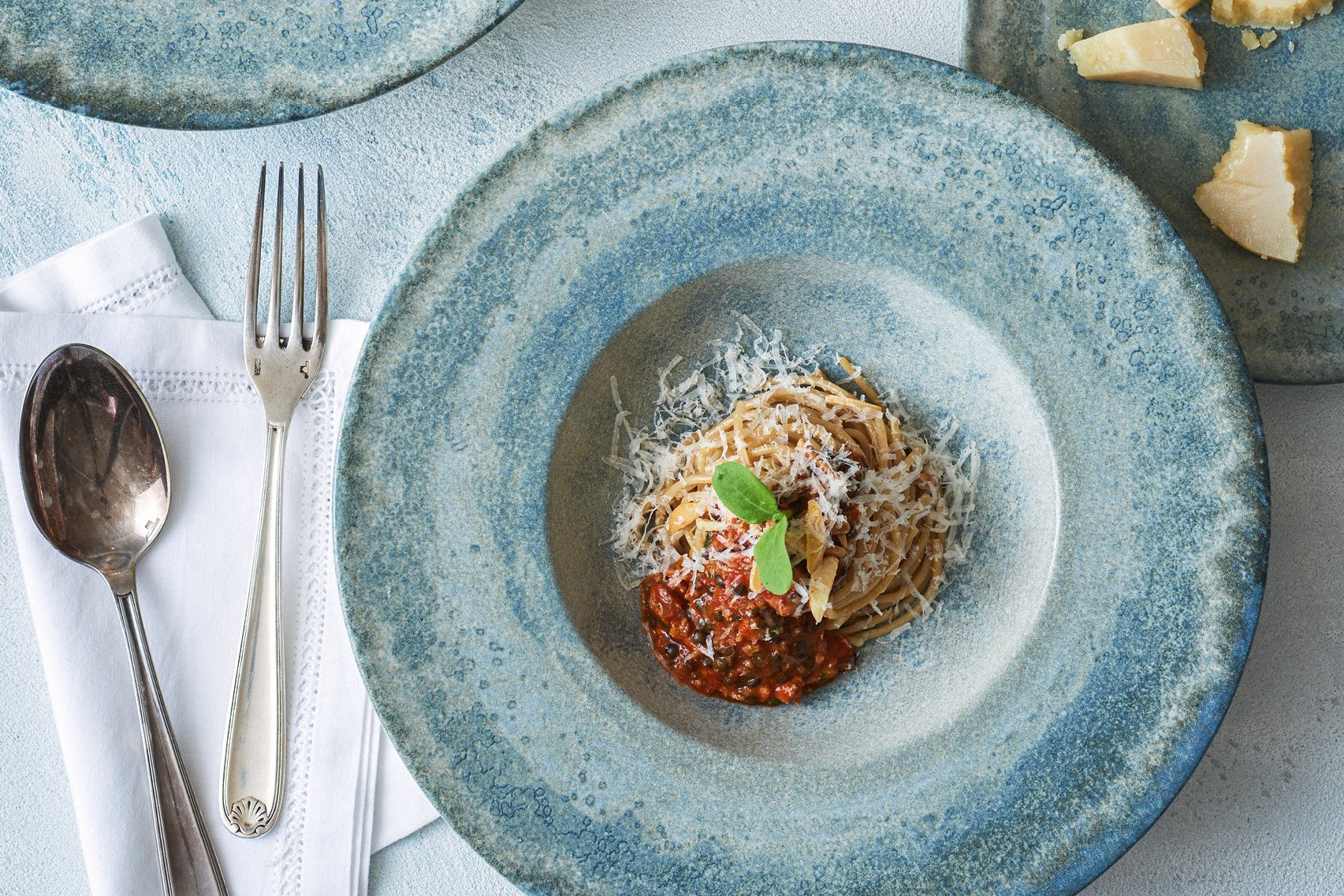Auf einem blauen tiefen Teller mit breiter Fahne sind Spaghetti mit Bolognesesoße angerichtet. Am Rand steht frischer Parmesan. Messer und Löffel im Shabby-Chic Look liegen links neben dem Teller auf einer weißen Spitzenserviette.