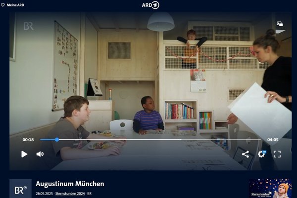 Ein Raum mit Holzverkleidung und 2 Ebenen ist zu sehen. An einem Tisch sitzen zwei Kinder. Rechts steht eine Frau mit großen Papierblättern in der Hand