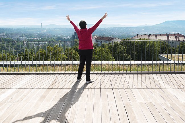 Eine Seniorin steht mit nach oben gestreckten Armen auf einer Terrasse