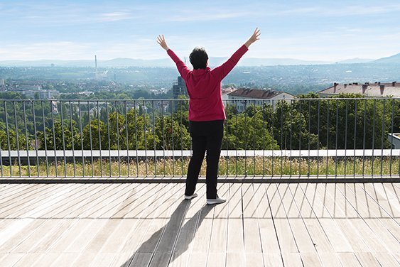 Eine Seniorin steht mit nach oben gestreckten Armen auf einer Terrasse