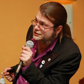 Ein Mann mit Brille und Mikrofon vor einem ockerfarbenen Hintergrund