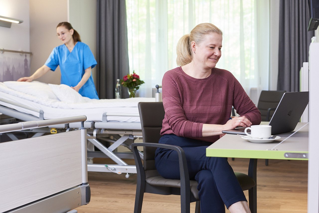Eine Frau sitzt an einem Tisch vor ihrem Laptop in einem Zimmer der Wahlleitungsstation und im Hintergrund macht eine Mitarbeiterin der Augustinum Klinik das Bett 