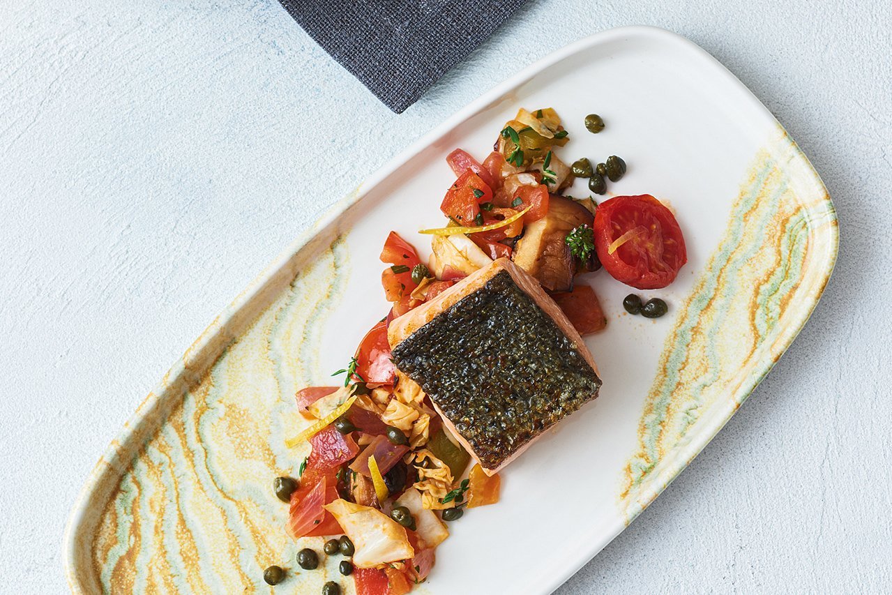 Auf einem weiß-gelben Teller mit gelb-grünen Wellen am Rand ist Tomaten-Pilz-Kraut-Gemüse längs angerichtet. Darauf ein kross gebratenes Stück Lachs. 