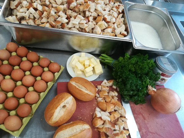 Brotstückchen, Semmeln, Zwiebeln, Butter, Eier und Petersilie liegen bereit zur Verarbeitung auf einem roten Brett  