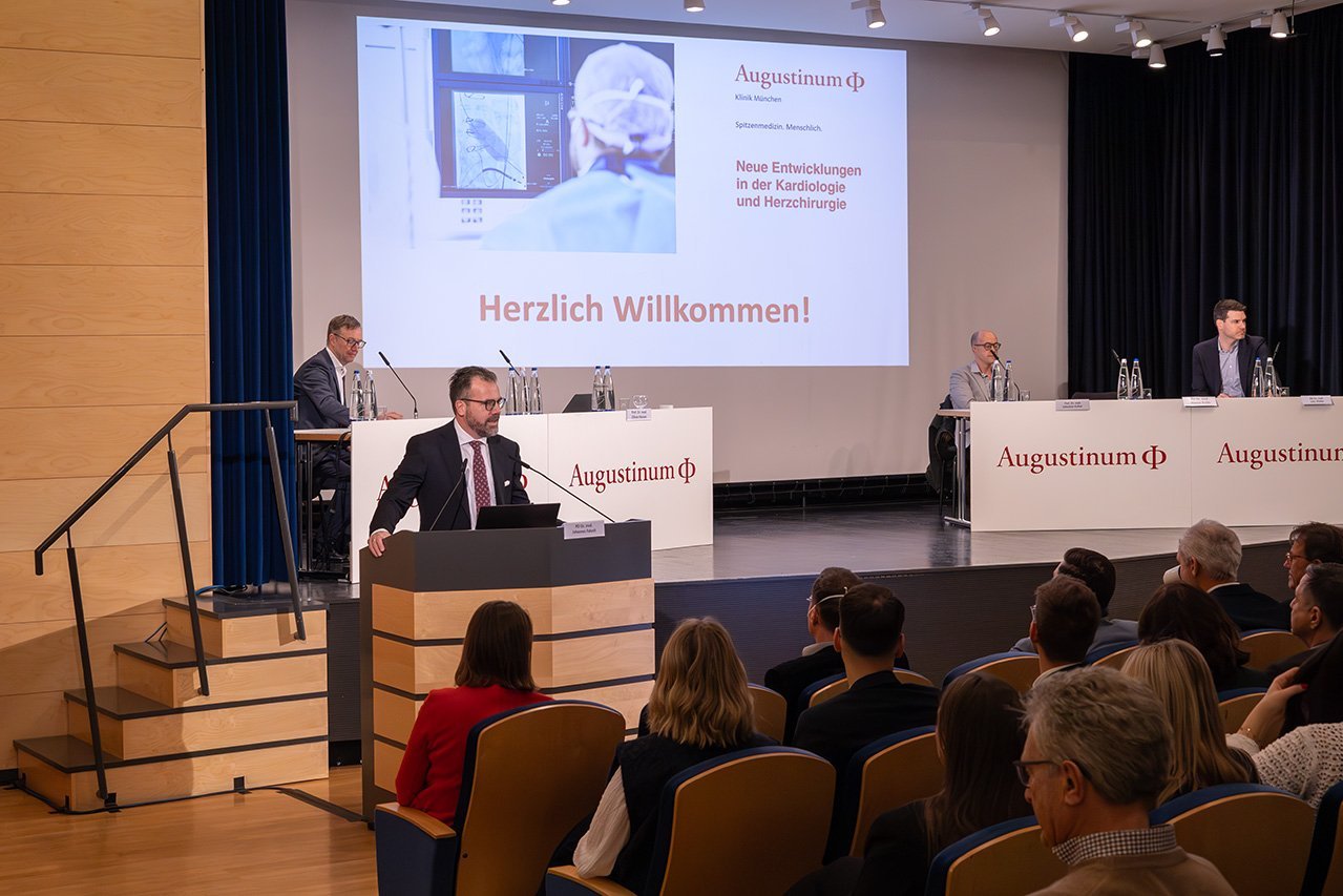 Dr. Husser, ein Chefarzt der Augustinum Klinik, steht an einem Rednerpult und spricht zum Publikum. Weitere Veranstalter sitzen auf der Bühne
