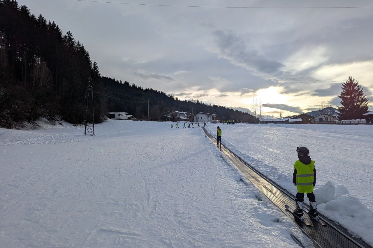 Eine große Schneefläche, durch die ein Weg führt, auf dem sich die Schüler*innen auf Skiern befinden