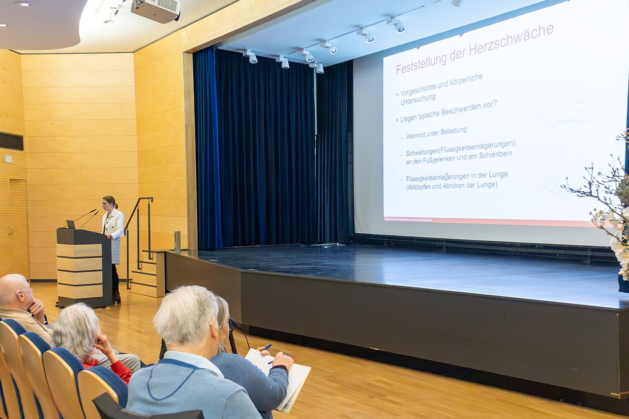 Besucher*innen betrachten die Präsentation im Theatersaal während eine Dozentin am Rednerpult einen Vortrag hält