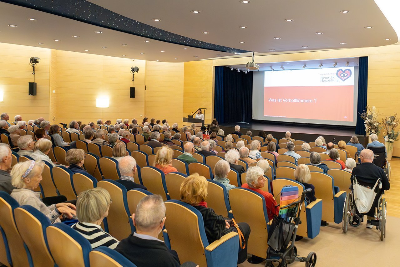 Viele Senior*innen sitzen in einem Theatersaal während eine Dozentin vor der Bühne einen Vortrag hält