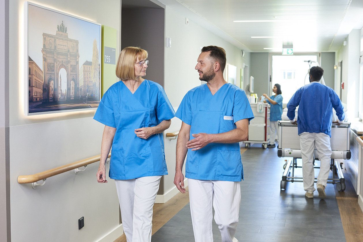Zwei Mitarbeiter*innen der Augustinum Klinik unterhalten sich im Flur einer Wahlleistungsstation, während im Hintergrund weitere Mitarbeiter*innen bei der Arbeit zu sehen sind