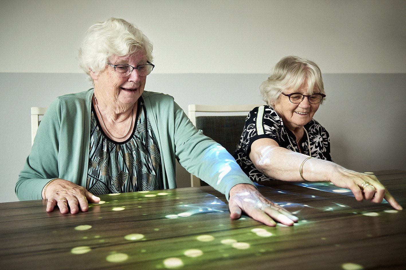 Zwei Seniorinnen sitzen am Tisch und spielen ein Spiel, das mithilfe einer Tovertafel auf den Tisch projiziert wurde