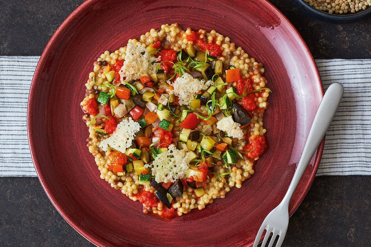 Auf einem tiefen Teller in kräftigem Rot ist breit gefächert eine Portion Pasta mit buntem Gemüse und Parmesanchips angerichtet. Am Tellerrand liegt eine Gabel.