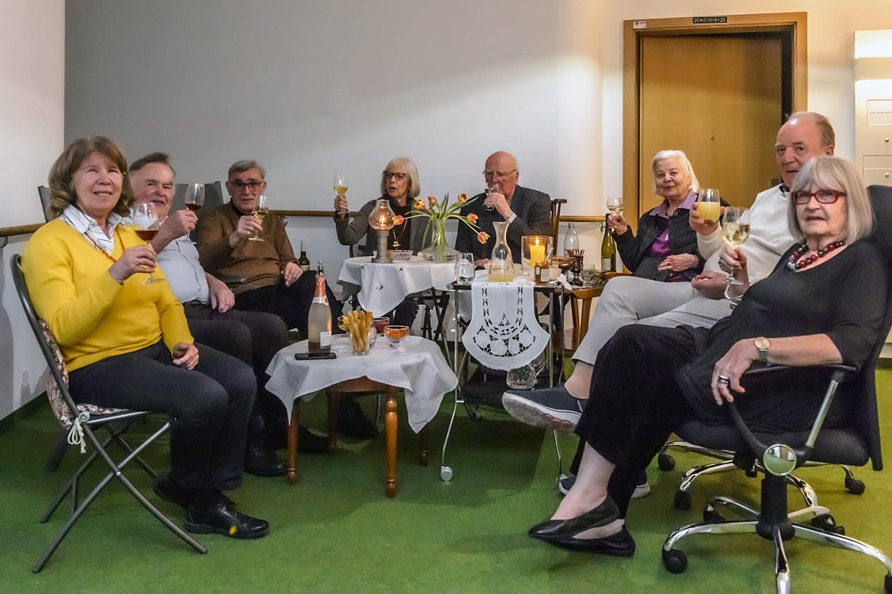 Acht Senior*innen sitzen an drei kleinen Partytischen und prosten zur Kamera