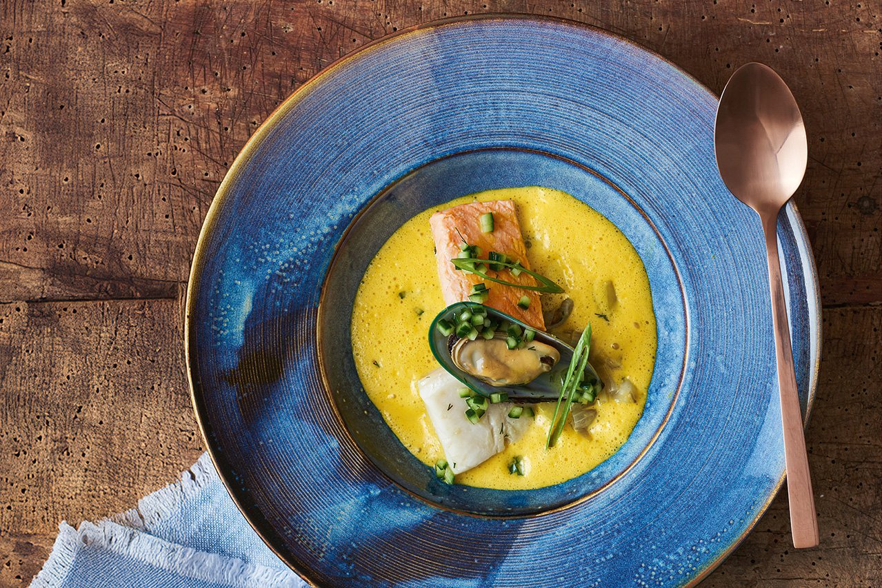 Ein tiefer dunkelblauer Teller mit breiter Fahne steht auf einem Holzhintergrund, am Rand liegt ein goldener Löffel. Darin befindet sich eine kräftig gelbe Suppe mit verschiedenen Fischteilen und einer Muschel in der Mitte. Frühlingszwiebeln und Gurkenstückchen sind als Deko darübergestreut.