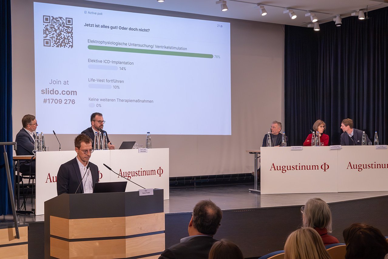 Fabian Friedel referiert am Rednerpult während das Veranstaltungsteam im Hintergrund auf der Bühne sitzt und eine Umfrage auf die Leinwand projiziert wird