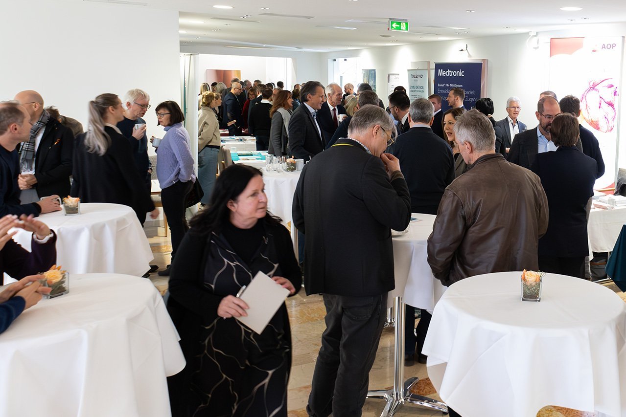 Viele Besucher*innen der Veranstaltung stehen in einem Raum mit kleinen weißen Tischen und unterhalten sich 