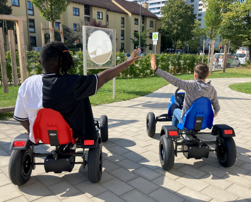Eine Aufnahme von hinten, bei der zwei Schüler auf Kettcars sitzen und deren Arm zur jeweils anderen Person strecken